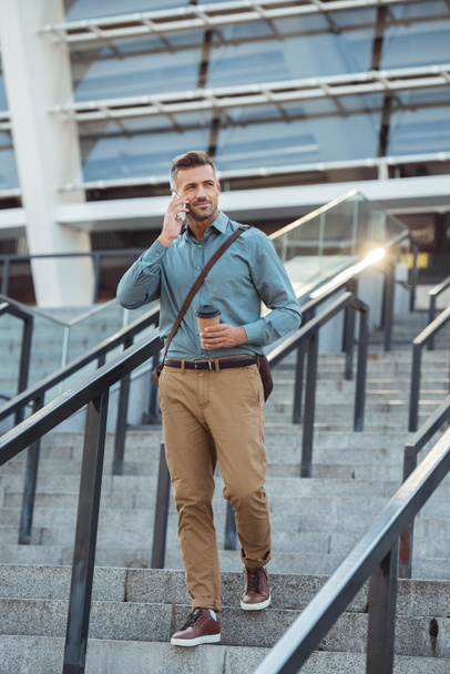handsome middle aged businessman holding coffee to go and talking by smartphone on stairs - Photo, Image