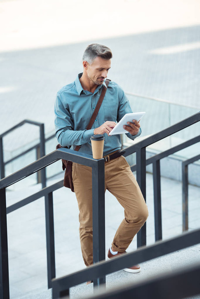 man using digital tablet while standing on stairs with coffee to go on railing - Foto, afbeelding