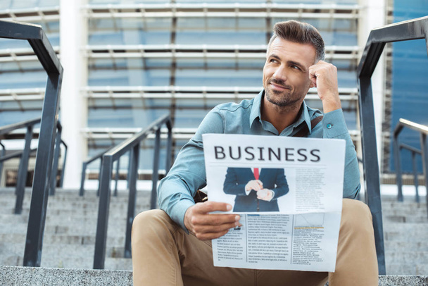 задумчивый улыбающийся мужчина держит деловую газету и смотрит в сторону, сидя на лестнице
 - Фото, изображение