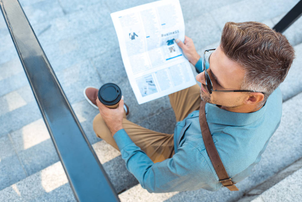 high angle view of man in sunglasses holding paper cup while sitting on stairs and reading newspaper  - Фото, изображение