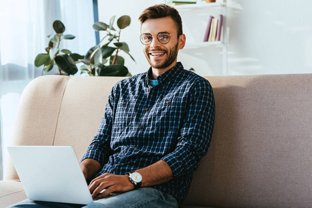 portrait of smiling man in eyeglasses with laptop sitting on sofa at home - Foto, Imagem