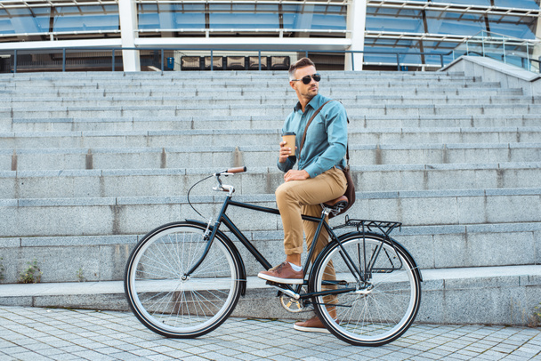 όμορφος άντρας με γυαλιά ηλίου που κάθεται στο ποδήλατο με καφέ να πάει και αναζητούν πάνω από τον ώμο στην οδό - Φωτογραφία, εικόνα