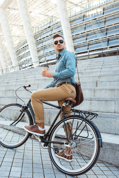 stijlvolle man in zonnebril op fiets zit ingedrukt te houden wegwerp koffiekopje - Foto, afbeelding