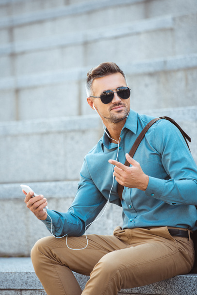 handsome middle aged man in earphones sitting on stairs and using smartphone   - Photo, Image