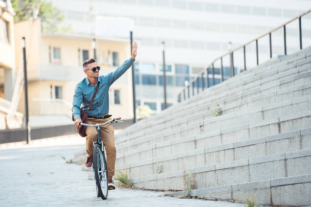 Glimlachende man in zonnebril fiets en zwaaien hand op straat - Foto, afbeelding