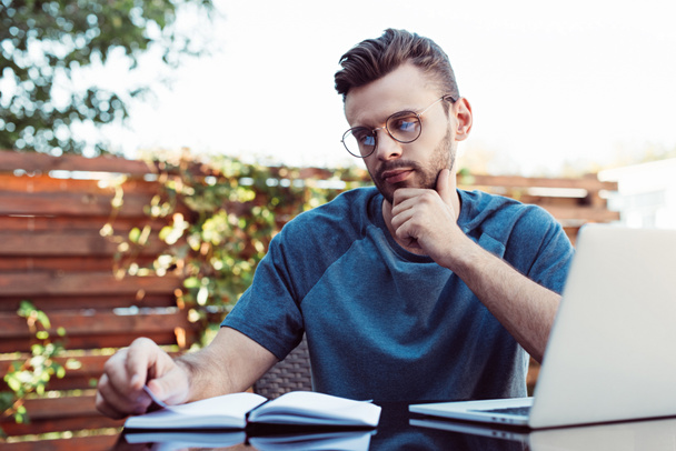 pensive handsome man taking part in webinar and looking at notebook in park - Fotografie, Obrázek