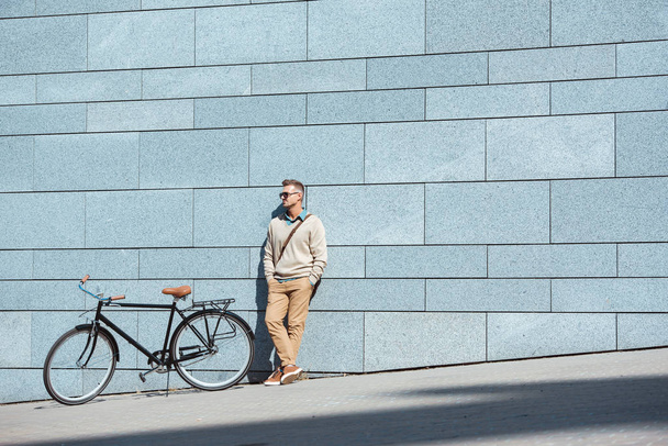 guapo hombre de mediana edad en gafas de sol de pie con las manos en los bolsillos cerca de la bicicleta y mirando hacia la calle
 - Foto, imagen