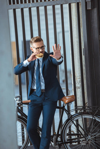 middle aged businessman gesturing by hand while sitting on bicycle and eating burger on street - Photo, Image