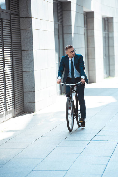 handsome middle aged businessman in suit and eyeglasses riding bicycle on street - Photo, image