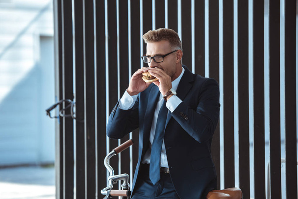middle aged businessman sitting on bicycle and eating hamburger - Zdjęcie, obraz
