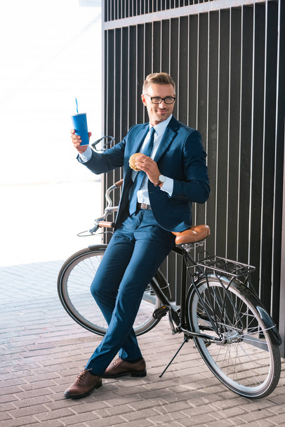 beau homme d'affaires souriant tenant tasse jetable et hamburger tout en étant assis sur le vélo
 - Photo, image