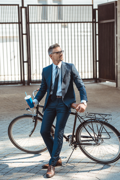 handsome smiling businessman holding paper cup and sitting on bicycle on street - Фото, изображение