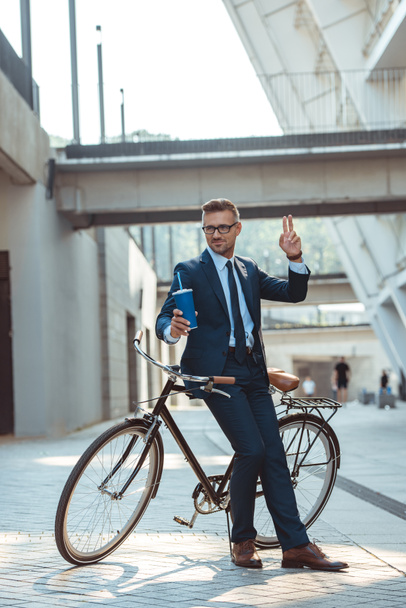 smiling businessman sitting on bicycle, holding paper cup and showing two fingers on street - Photo, Image