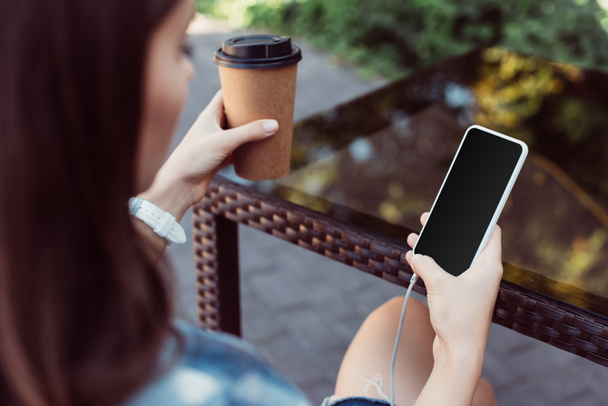 Frau benutzt Smartphone mit leerem Bildschirm am Tisch und hält Kaffee in Pappbecher - Foto, Bild
