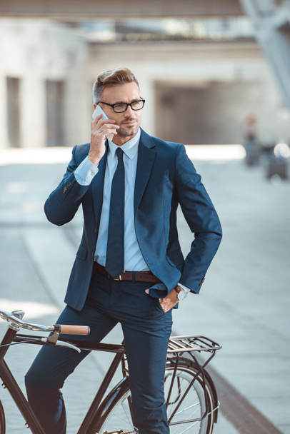 handsome middle aged businessman in formal wear and eyeglasses sitting on bicycle and talking by smartphone - Foto, afbeelding