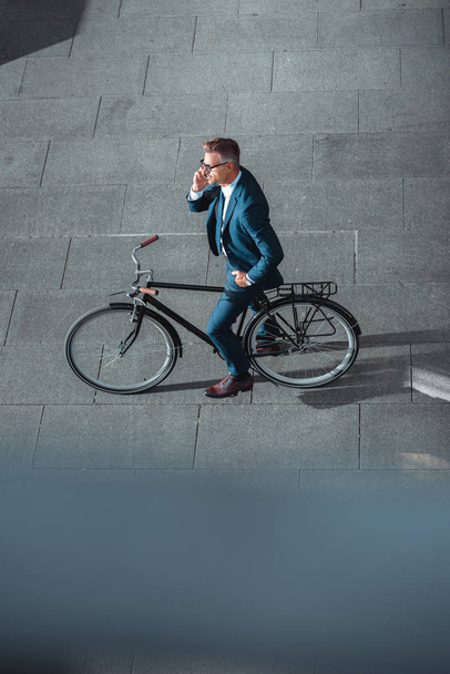 high angle view of businessman in formal wear talking by smartphone while standing with bicycle on street  - Photo, Image