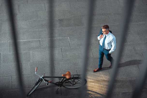 επιλεκτική εστίαση του επιχειρηματία κρατώντας κοστούμι σακάκι και κοιτάζοντας ποδηλάτων στην οδό    - Φωτογραφία, εικόνα