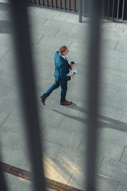 selective focus of businessman with coffee to go and newspaper walking on street  - 写真・画像