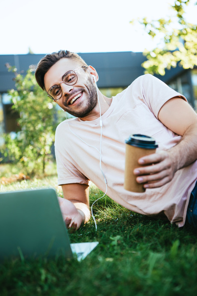 smiling handsome man taking part in webinar and lying on grass in park - Photo, Image