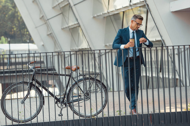 επιτυχημένος μέσης ηλικίας επιχειρηματίας στην επίσημη ένδυση κρατώντας χαρτί Κύπελλο και τον έλεγχο Ρολόι χεριού ενώ στέκεται κοντά στο ποδήλατο - Φωτογραφία, εικόνα