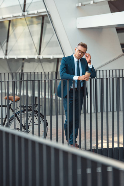 businessman in formal wear holding coffee to go and adjusting eyeglasses while standing near bike on street - Zdjęcie, obraz