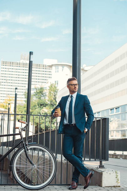 handsome businessman holding coffee to go and looking away while standing near bicycle on street - Zdjęcie, obraz