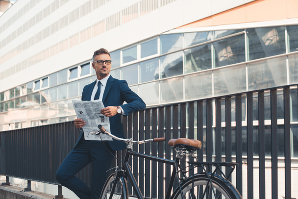 successful businessman in formal wear holding newspaper and looking away while standing with bicycle on street - Foto, afbeelding