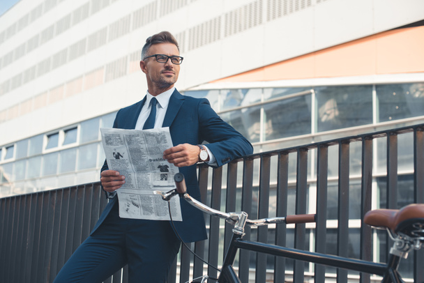 Businessman in suit and eyeglasses holding newspaper and looking away while standing with bike on street - Photo, Image