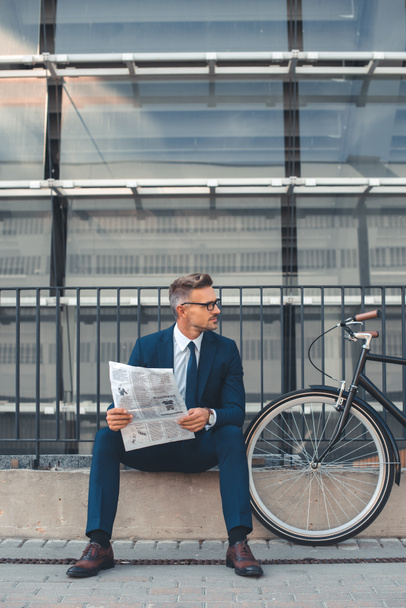 Geschäftsmann mittleren Alters hält Zeitung in der Hand und schaut weg, während er neben dem Fahrrad auf der Straße sitzt  - Foto, Bild