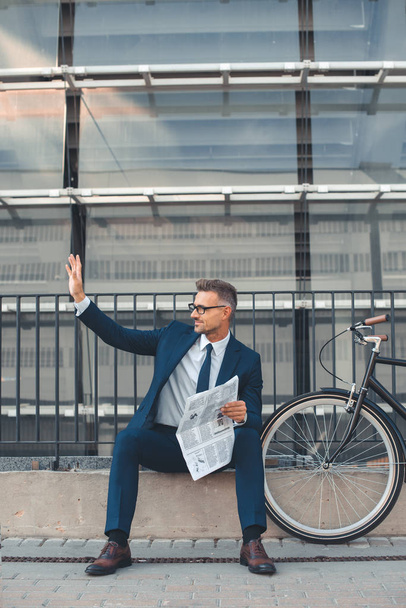 Geschäftsmann mittleren Alters hält Zeitung in der Hand und winkt mit der Hand, während er neben dem Fahrrad auf der Straße sitzt  - Foto, Bild