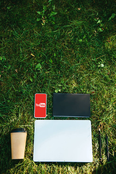 top view of smartphone with youtube appliance, laptop and coffee in paper cup on green grass in park - Foto, afbeelding