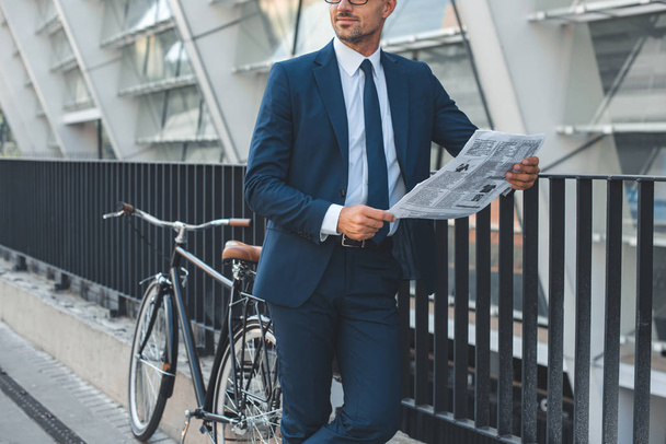 обрізаний знімок бізнесмена в офіційному одязі, що тримає газету, стоячи з велосипедом на вулиці
 - Фото, зображення