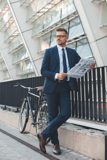 schöner Geschäftsmann mit Brille hält Zeitung in der Hand und schaut weg, während er mit dem Fahrrad auf der Straße steht - Foto, Bild
