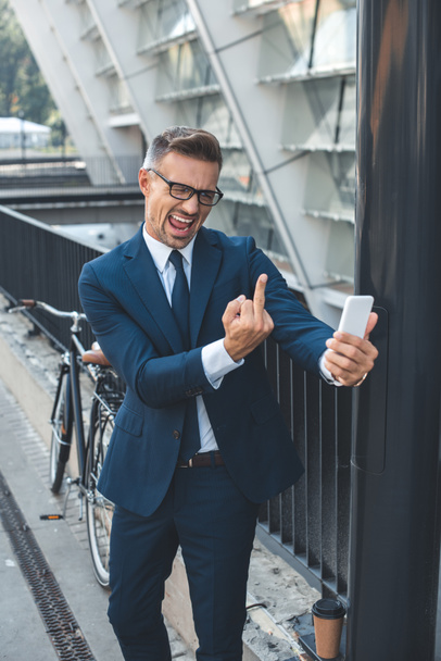 emotional middle aged businessman in formal wear and eyeglasses holding smartphone and showing middle finger - Фото, изображение