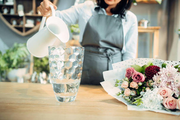 Female florist pours water into a vase in flower shop. Floristry service, floristic business - 写真・画像