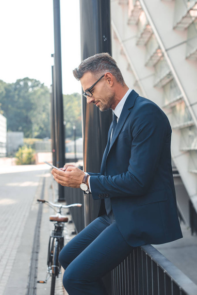 Seitenansicht eines Geschäftsmannes in Anzug und Brille mit Smartphone auf der Straße - Foto, Bild