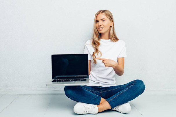 Portrait of beautiful girl wearing white t-shirt showing blank laptop screen sitting on floor isolated on grey background - Photo, image
