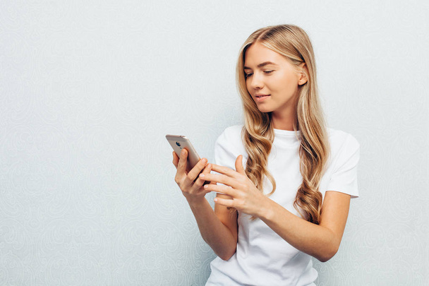 Young beautiful girl dressed in white shirt, holding the phone and staring at the screen, sitting on the floor cross-legged, isolated on grey background - Photo, image