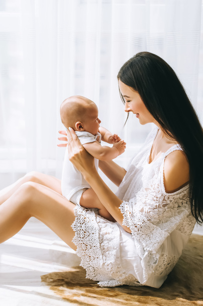 attractive young mother sitting on floor near window with her adorable little child at home - Foto, Imagen