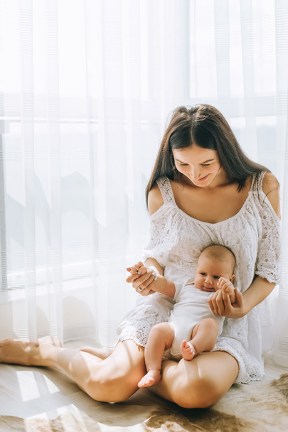 happy young mother sitting on floor near window with her adorable little child at home - Φωτογραφία, εικόνα