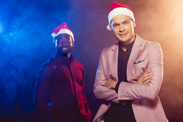 handsome man in pink jacket and santa hat standing near african american friend on black with smoke and backlit  - Fotografie, Obrázek