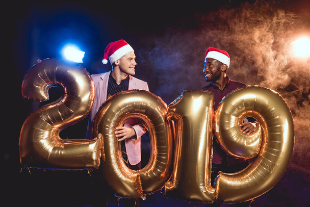 monikulttuurinen ystävät santa hatut katselevat toisiaan pitäen 2019 uusi vuosi kultainen ilmapalloja savua ja taustavalaistu
  - Valokuva, kuva