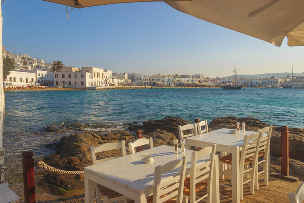 Chairs with tables in typical Greek restaurant  of Mykonos old town, Cyclades island, Greece. Panoramic view of the marina, the sandy beach and the characteristic white houses. - Fotoğraf, Görsel