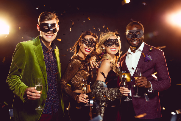 glamorous multiethnic friends in carnival masks holding champagne glasses and celebrating new year on party - Photo, Image