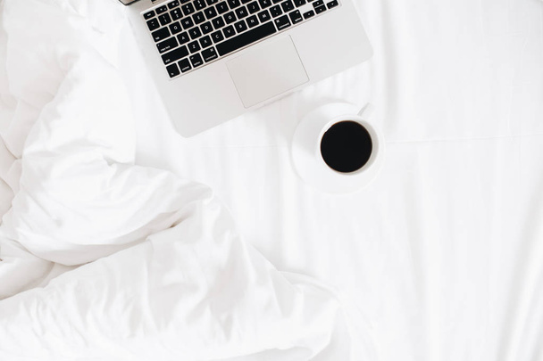 Ordenador portátil y café en la cama con ropa de cama blanca. blogger freelance workspace flat lay. Vista superior fondo mínimo
. - Foto, Imagen