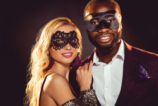 glamorous multiethnic smiling couple in carnival masks for new year party - Photo, Image