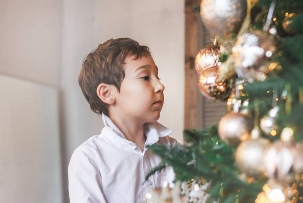 Cute boy 8 years old in a white shirt adorns the Christmas tree - Foto, Imagen