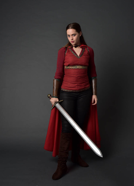 po celé délce portrét bruneta dívka, která nosí červený středověký kostým a plášť. stojící pozice drží meč na šedé studio pozadí. - Fotografie, Obrázek