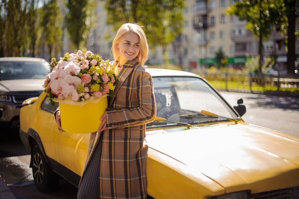 Дівчина в осінньому пальто тримає в руках красиву жовту коробку з різних квітів, що стоять біля таксі
 - Фото, зображення