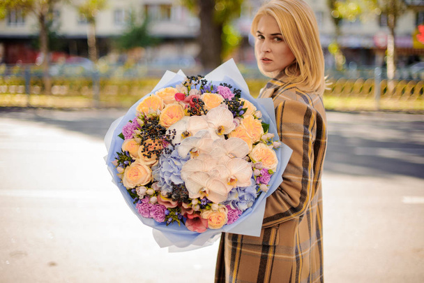Girl in the autumn coat holding in her hands a beautiful bouquet of different flowers in violet paper - Fotó, kép